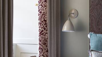 A hotel room with a bed, a bedside table with a rotary phone, a wall-mounted lamp, and patterned curtains.