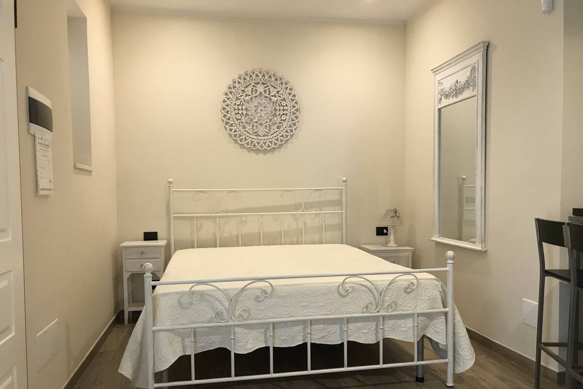 Ein Schlafzimmer mit einem weißen Metallbett, einem Nachttisch, einem Wanddekor und einem Spiegel.