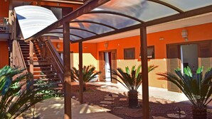 Un'area esterna coperta con una scala che conduce a un piano superiore, circondata da palme e un edificio con pareti arancioni e persiane in legno.