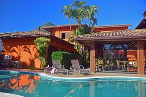 Un área de piscina con sillones de descanso y un comedor, rodeado de palmeras y un edificio con techo de tejas.