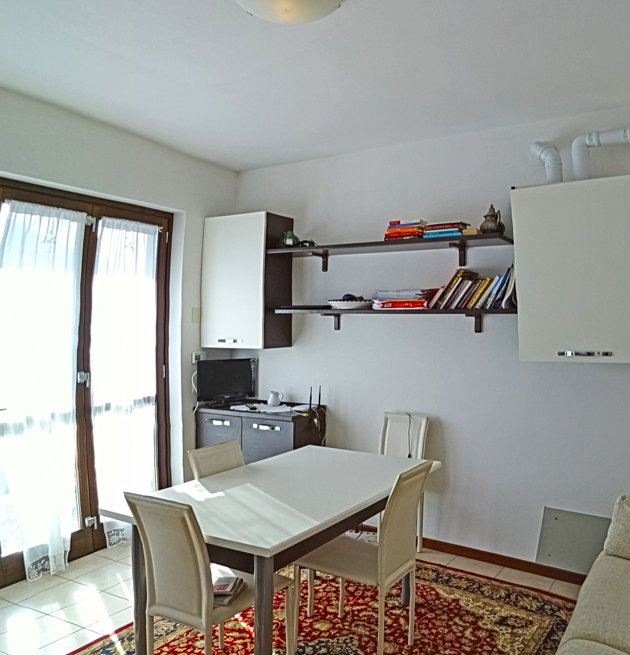 Un comedor con mesa y sillas blancas, una cocina con estufa y alacenas, y una estantería con libros y objetos decorativos.