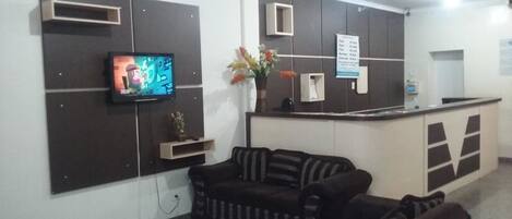 Zona de recepción con televisor de pantalla plana montado en la pared, mostrador de recepción, sofá y un jarrón con flores.