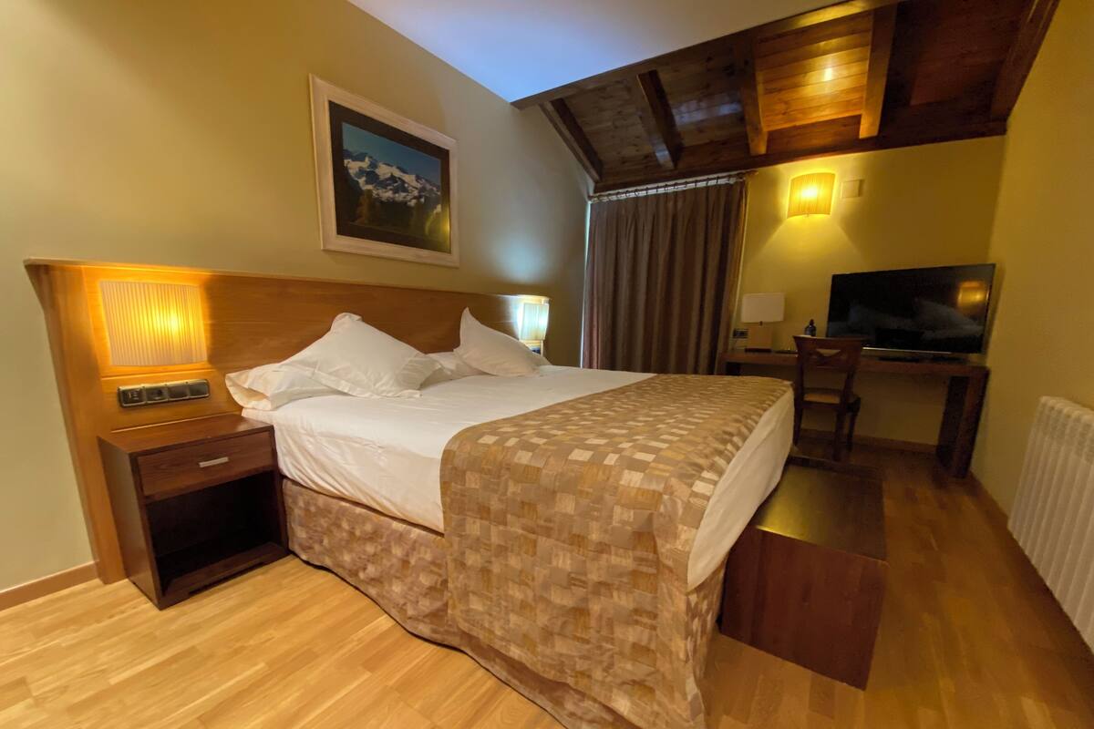 A hotel room with a large bed, a wooden headboard, a bedside table with a lamp, a TV, and a painting on the wall.