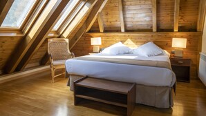 Een slaapkamer met een schuine kap, houten balken, een bed met witte lakens, twee nachtkastjes met lampen en een stoel.