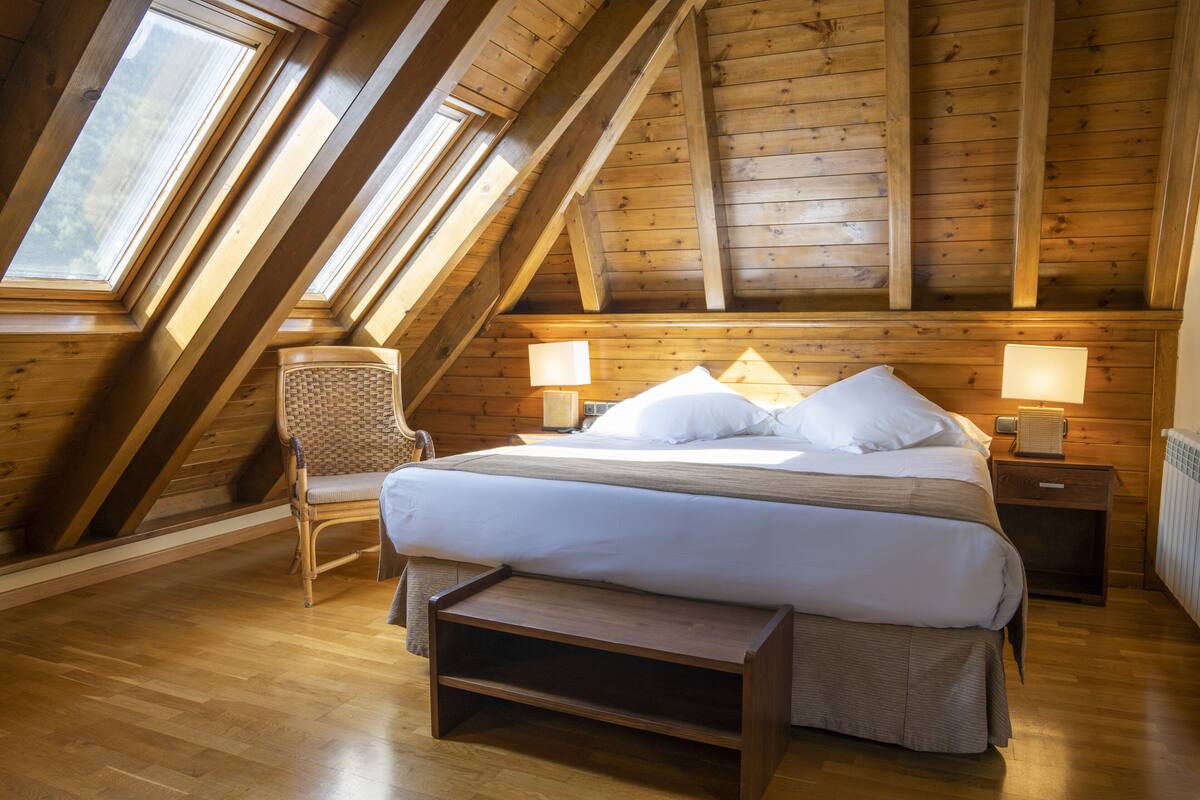 A bedroom with a sloped ceiling, wooden beams, a bed with white linens, two bedside tables with lamps, and a chair.
