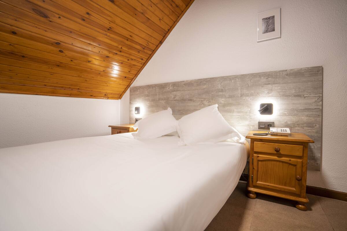 A bedroom with a wooden ceiling, a bed with white linens, a nightstand with a lamp, and a wall-mounted light.