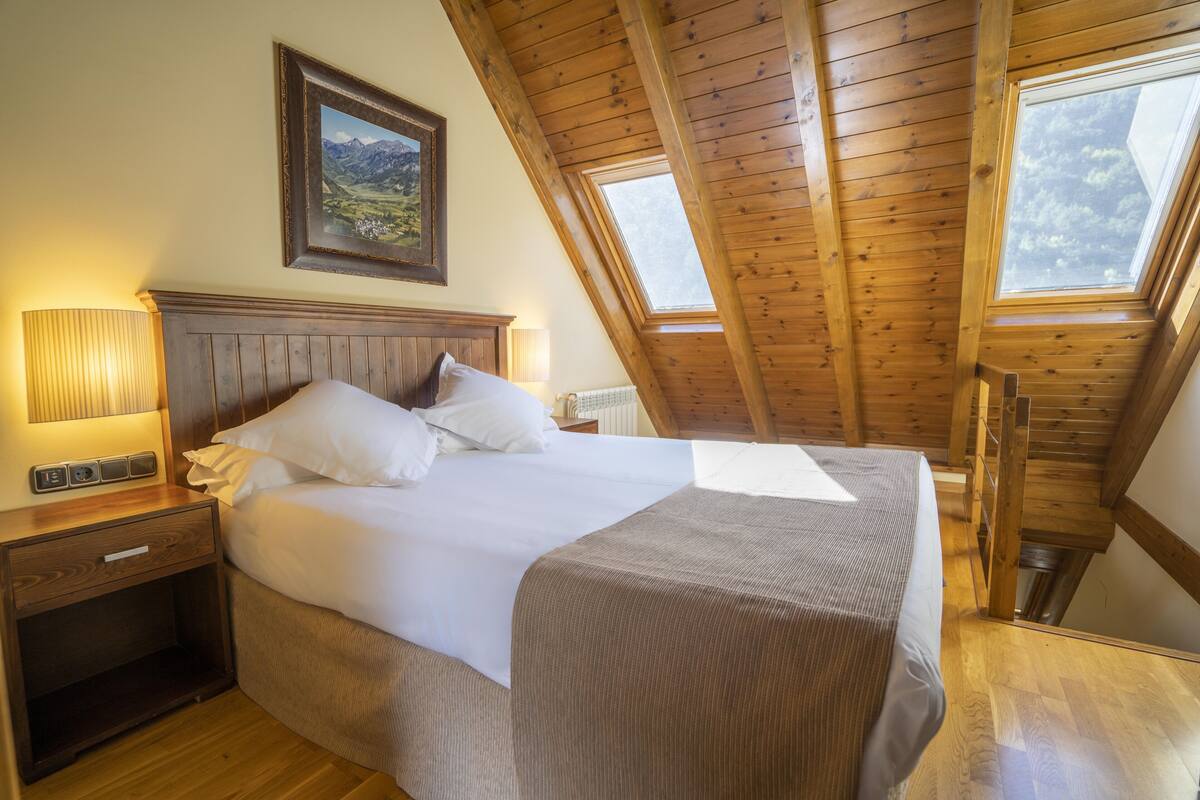 A bedroom with a wooden ceiling, a bed with white linens, a nightstand, and a wall-mounted lamp.
