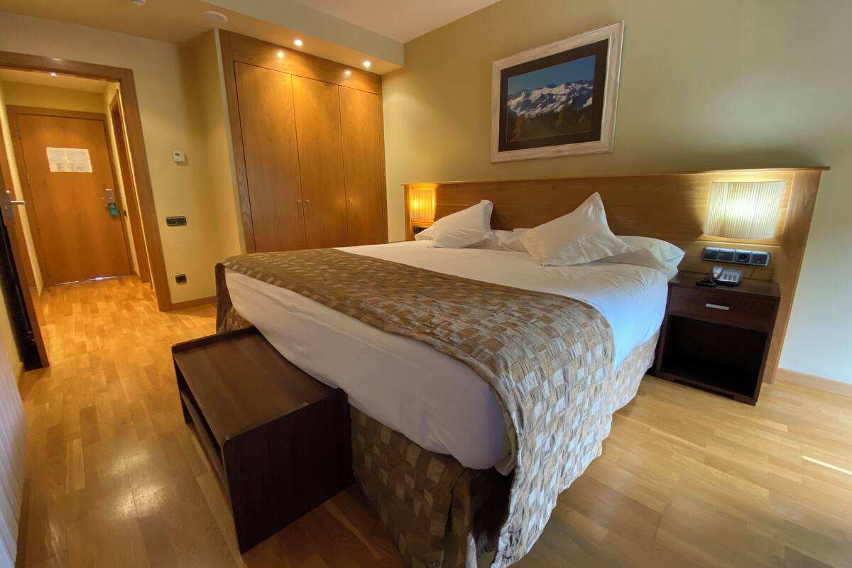 A hotel room with a large bed, a wooden wardrobe, a nightstand with a lamp, and a framed picture on the wall.