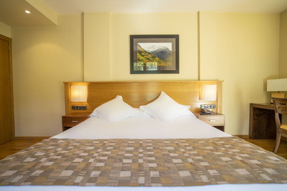 A hotel room with a large bed, wooden headboard, two bedside tables with lamps, a desk, and a chair.