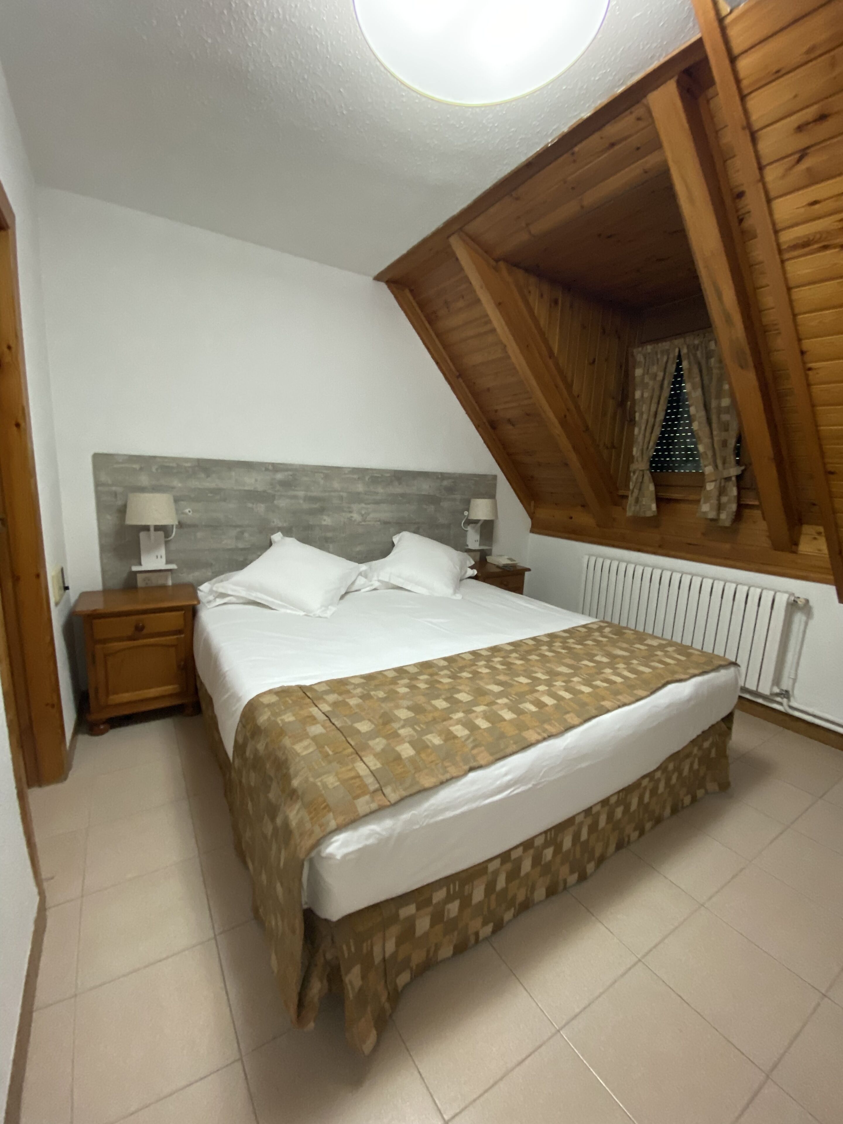 A bedroom with a bed, two bedside tables, a window with curtains, and a sloped ceiling.