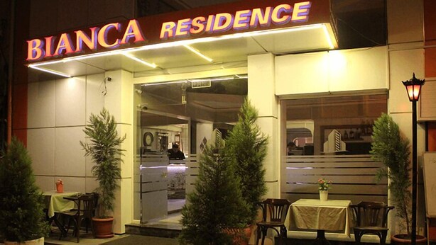 Das Außengelände des Bianca Residence mit einer Neonreklame, Sitzbereich im Freien und Topfpflanzen.