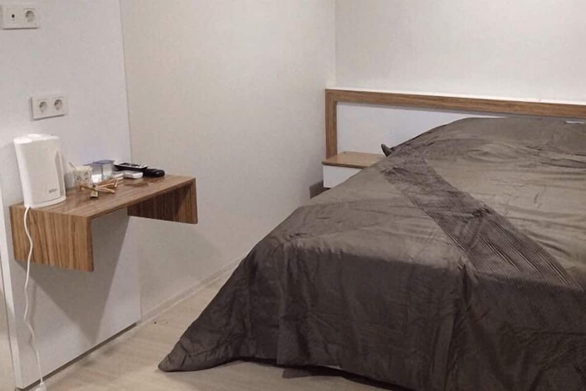 A bedroom with a bed covered in a grey blanket, a bedside table with a lamp, and a wall socket.