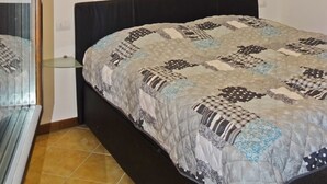 Une chambre à coucher avec un lit, une fenêtre avec des stores, un luminaire au plafond et un sol carrelé.