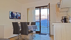 Un coin repas avec une table et des chaises, un téléviseur fixé au mur et une vue sur les montagnes par une porte vitrée.