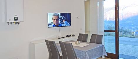 A modern dining area with a flat-screen TV mounted on the wall, a white dining table with four chairs, and a view of a mountainous landscape through a large sliding glass door.