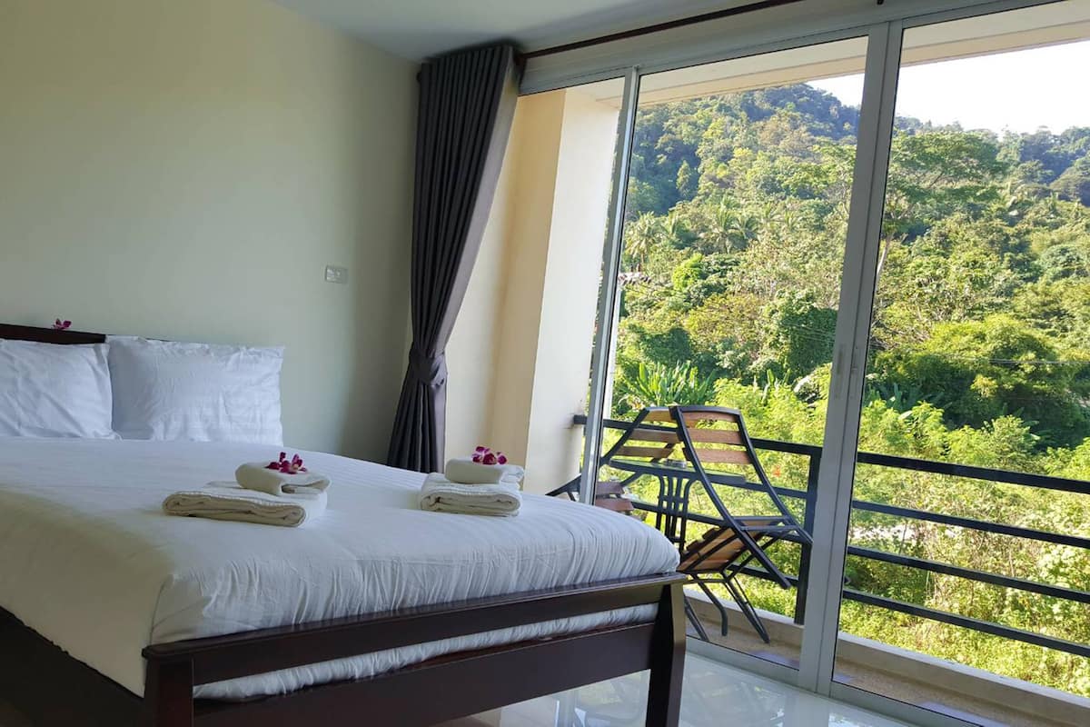Habitación de hotel con una cama grande, un balcón con vistas a zona boscosa y una silla.
