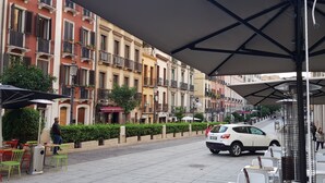 A street with outdoor seating, a white car, and multi-story buildings with balconies. - The Good Place (Cagliari)