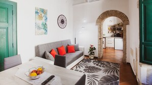 A cozy living room with a sofa, a dining table with fruit, a clock, and a kitchen visible through an archway. - The Good Place (Cagliari)