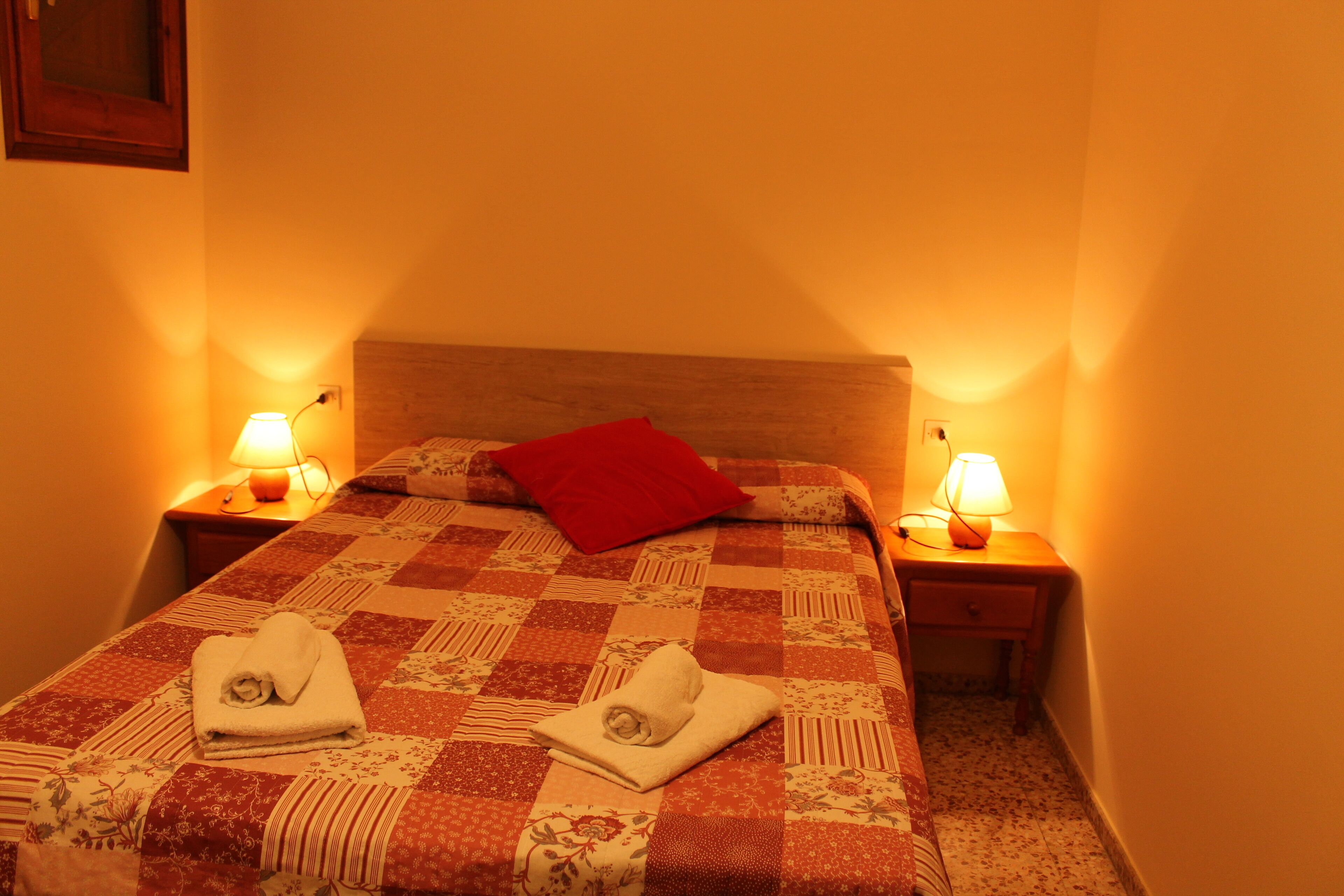 A neatly made bed with a red pillow, two bedside lamps, and two folded towels on the bed.