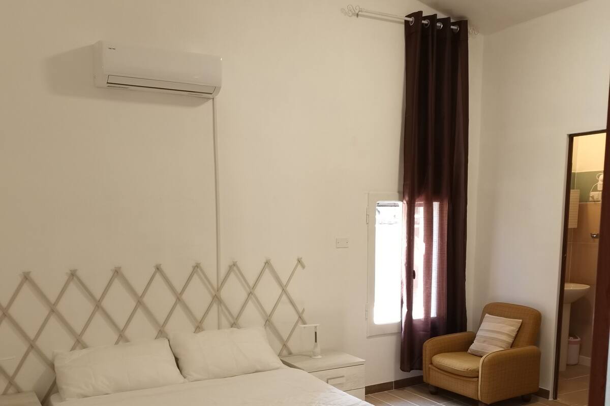 A bedroom with a bed, a nightstand, a chair, an air conditioning unit, and a window with curtains.