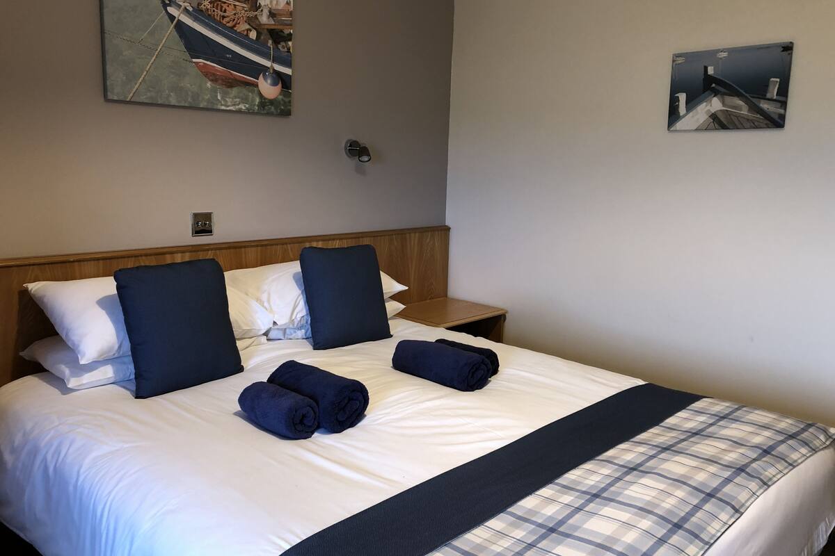 A neatly made bed with white and dark blue linens, a plaid blanket, and two rolled towels. There are two paintings on the walls, one depicting a boat and the other a building.