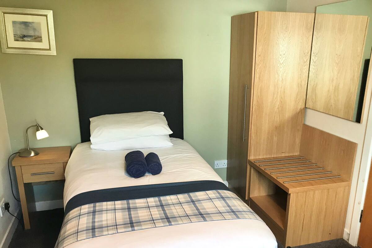 A neatly made bed with a black headboard, a bedside table with a lamp, and a wooden wardrobe with a mirror.