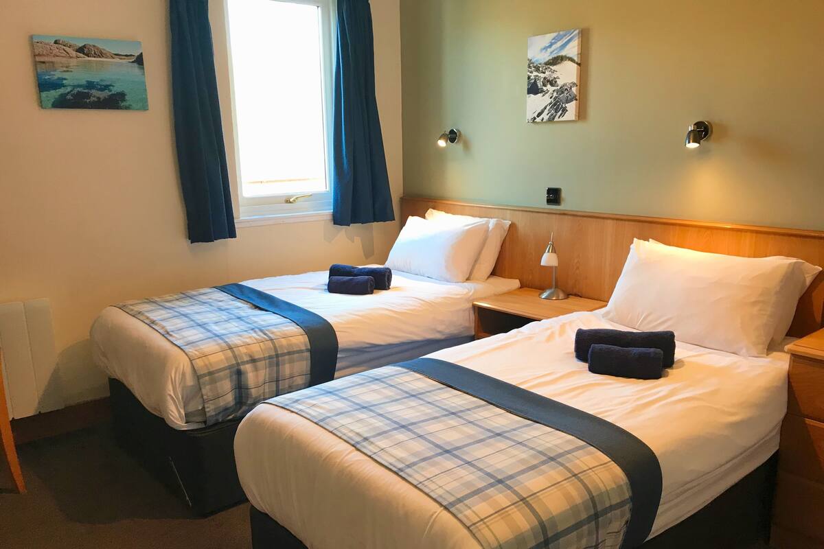 A double room with two beds, each with a plaid bedspread and pillows, a nightstand with a lamp, and a window with curtains.