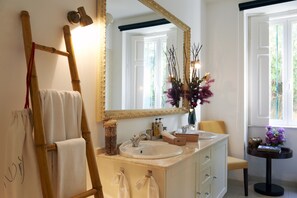 A bathroom with a large mirror, two sinks, a towel rack, and a small table with a vase of flowers. - Lucky's Guest House (Cascais)