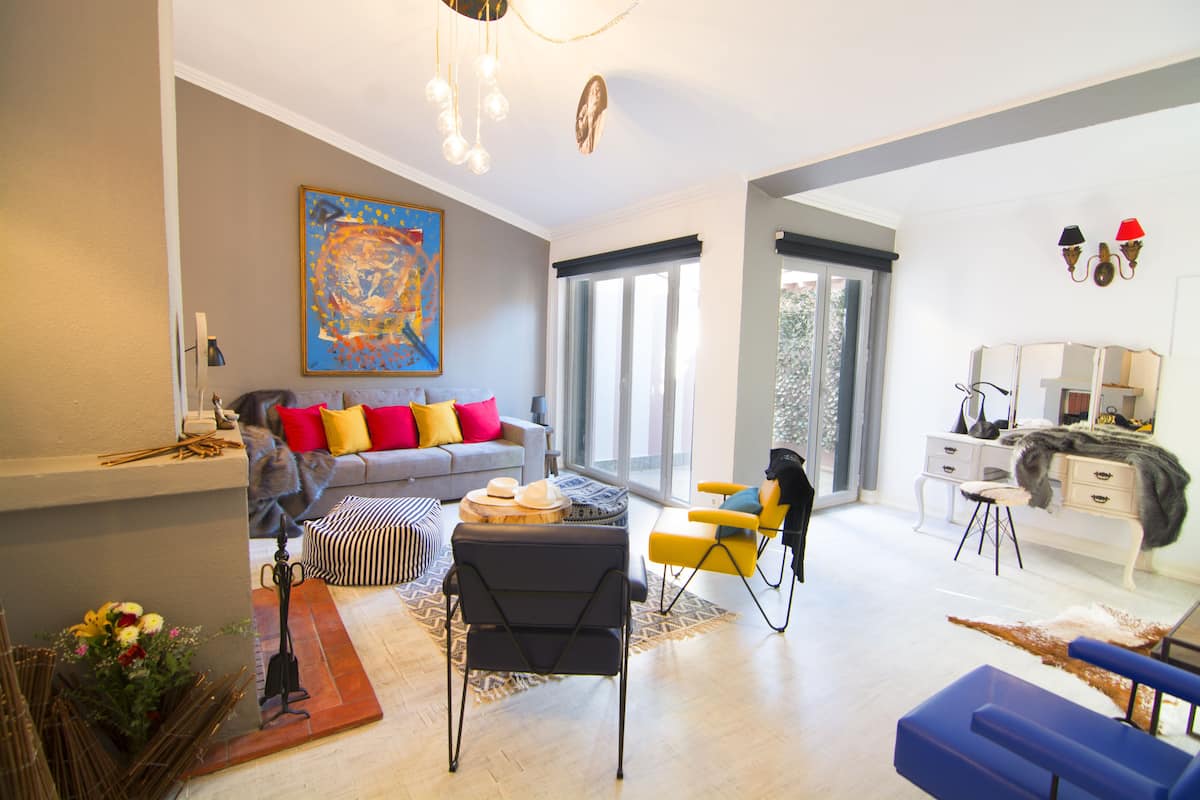 Una sala de estar moderna con un sofá gris, cojines coloridos, una silla negra y una silla amarilla. Hay un cuadro en la pared y una ventana con persianas.
