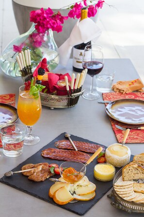 A table set with a variety of food items including cheese, bread, and a glass of wine, with a vase of flowers in the background. - Lucky's Guest House (Cascais)