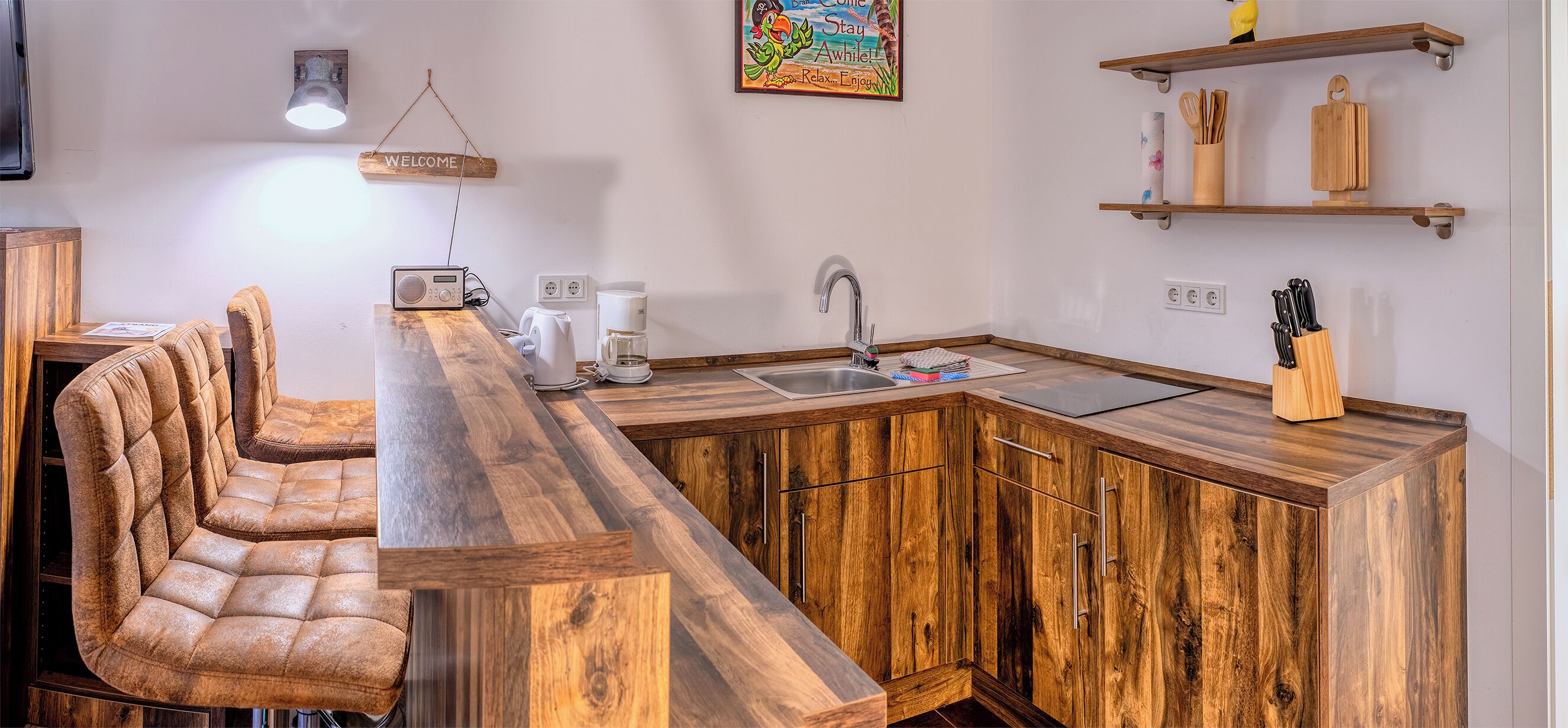 Une cuisine avec un bar en bois, un évier et une machine à café. On y trouve des chaises et une table sur laquelle est posée une pancarte indiquant « BIENVENUE ».