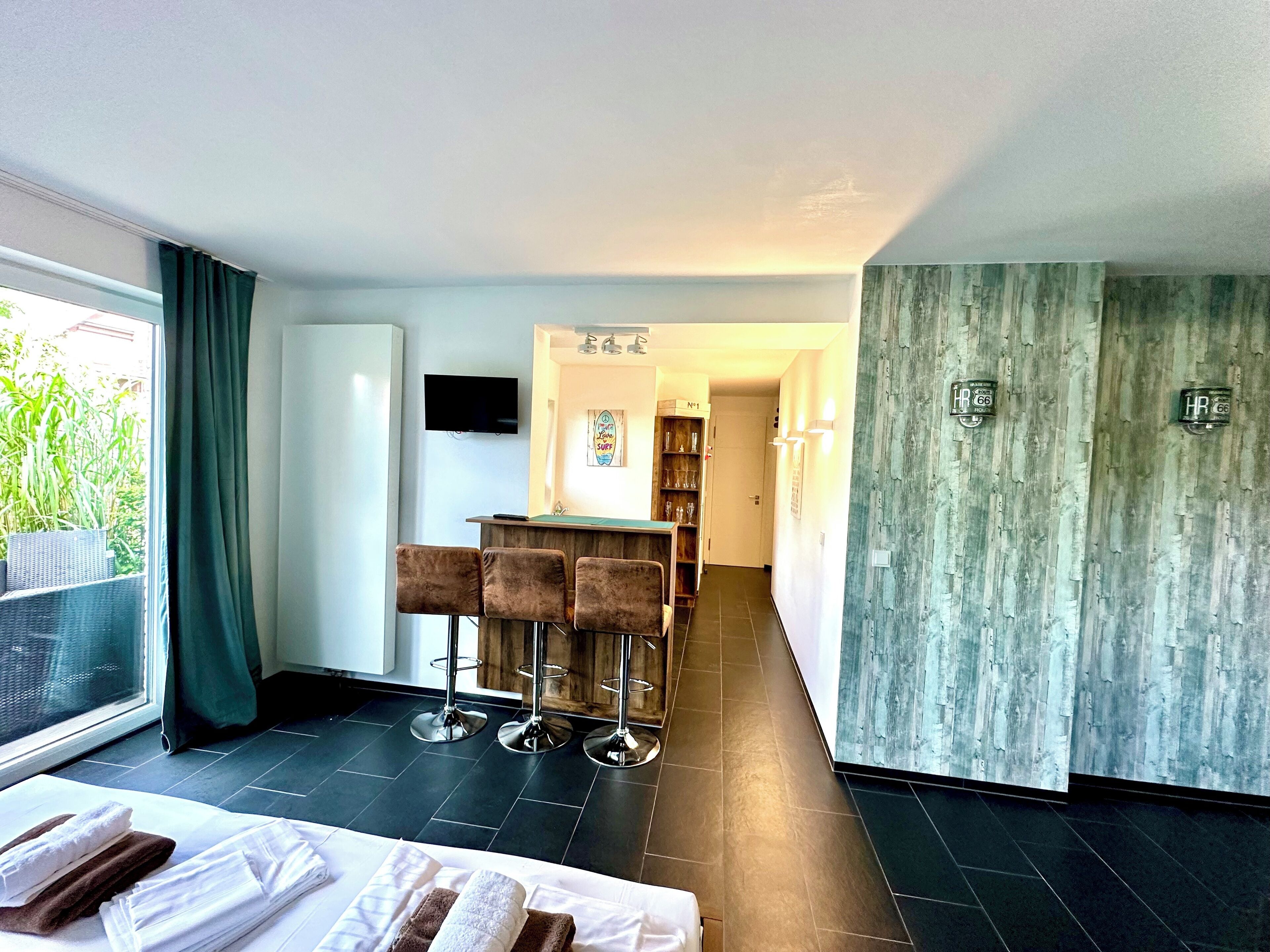 A modern hotel room with a bed, a dining area with bar stools, a TV mounted on the wall, and a view of greenery outside.