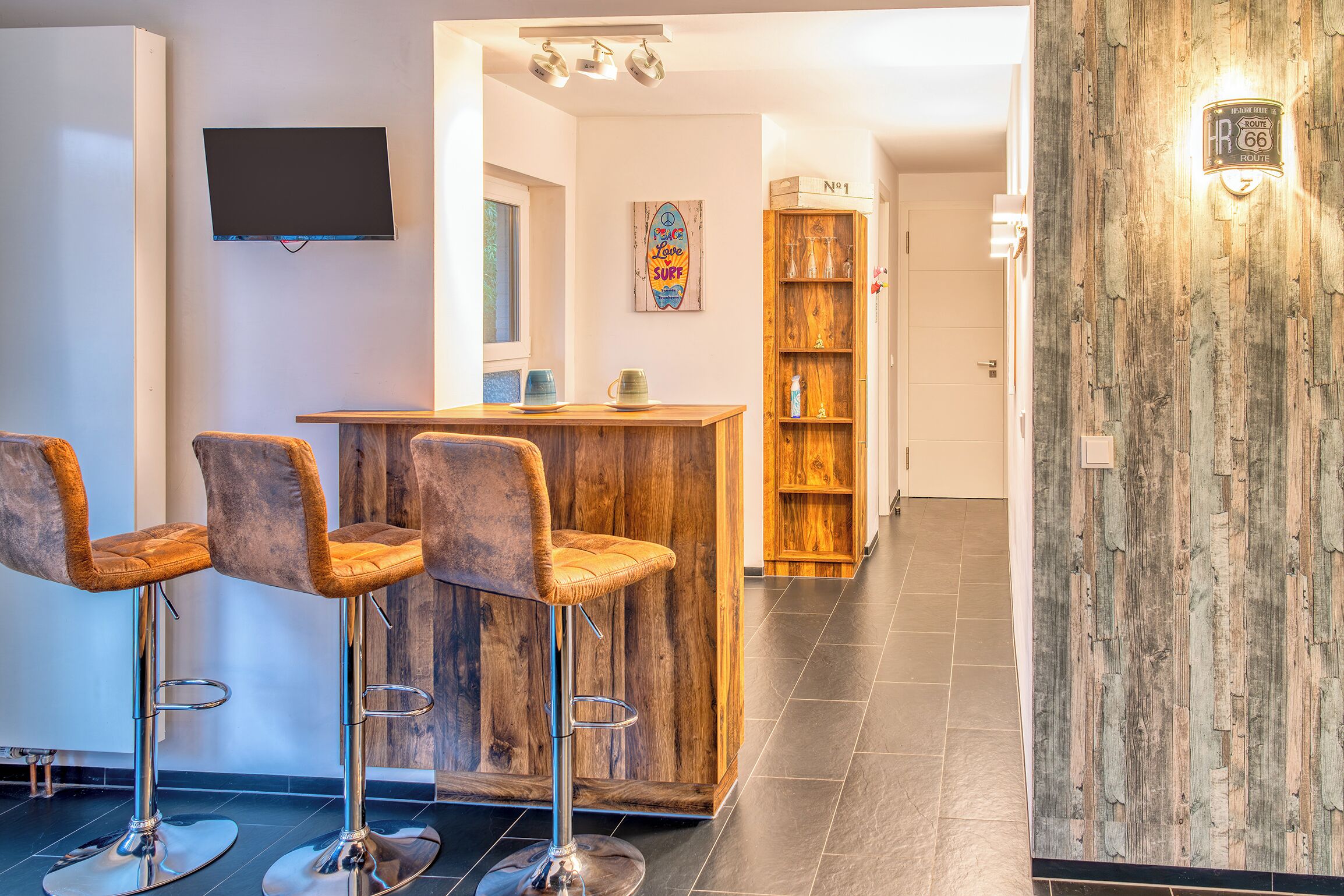 A bar area with two bar stools, a wooden bar counter, a television mounted on the wall, and a surfboard artwork.