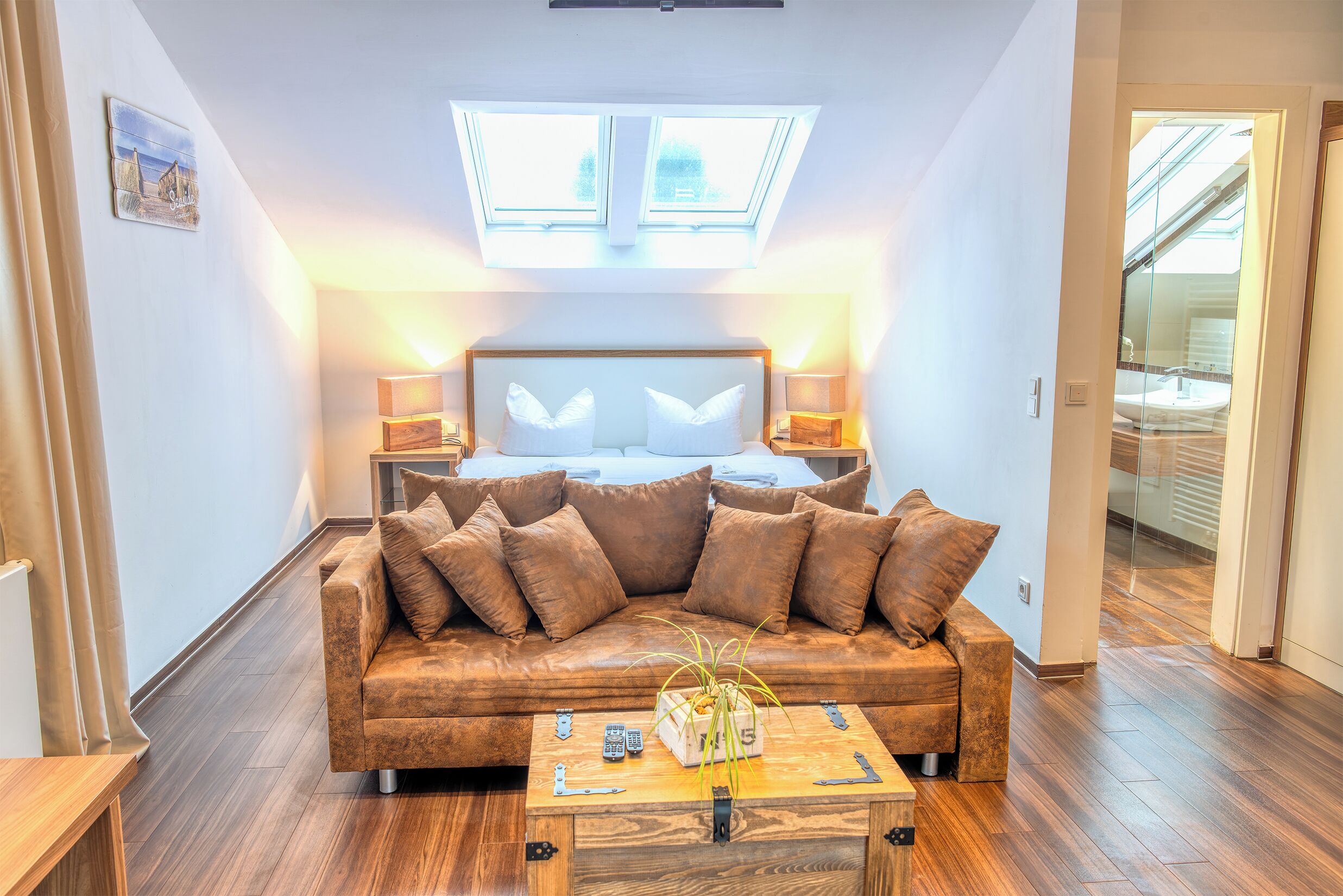 Une chambre à coucher moderne avec un canapé en cuir marron, une table basse en bois agrémentée d’une plante, et une lucarne au-dessus du lit.