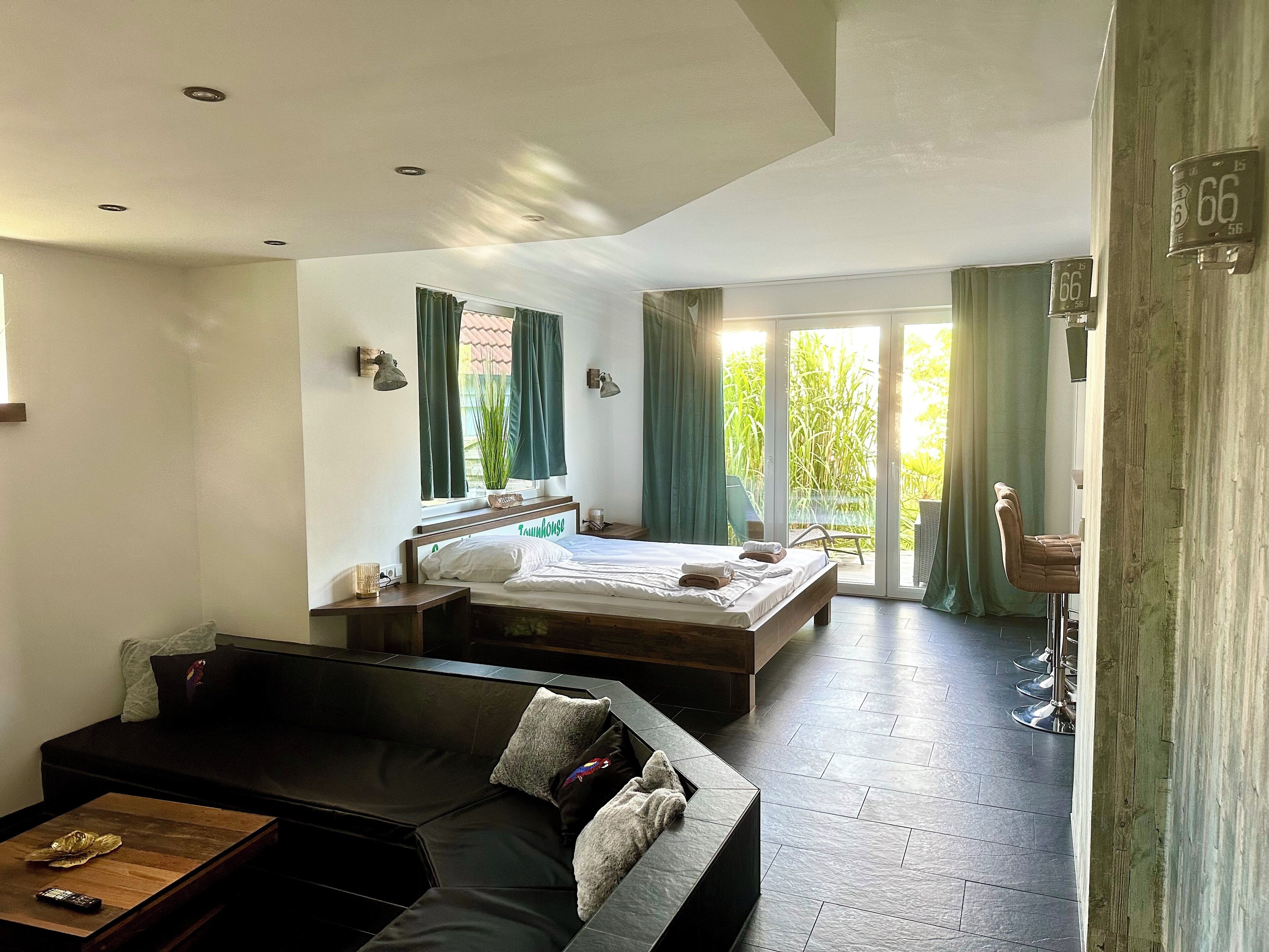 A modern hotel room with a large bed, a sofa, and a dining area with a view of greenery.
