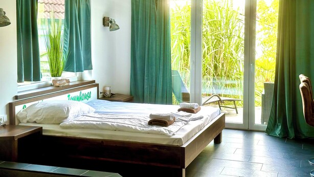 Een slaapkamer met een bed, een nachtkastje, een lamp en uitzicht op groen buiten het raam.