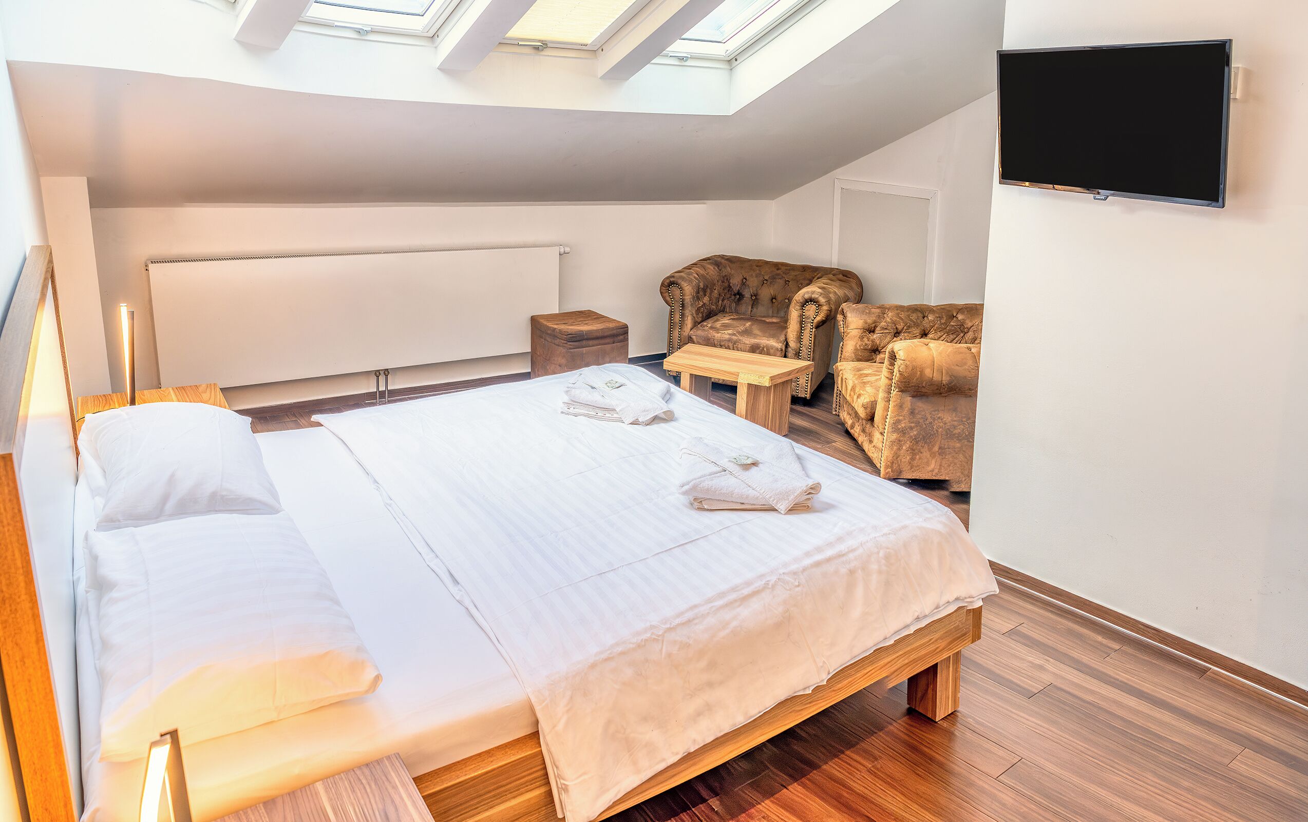 Un dormitorio con cama, mesita de noche de madera, una silla, televisor y tragaluz.