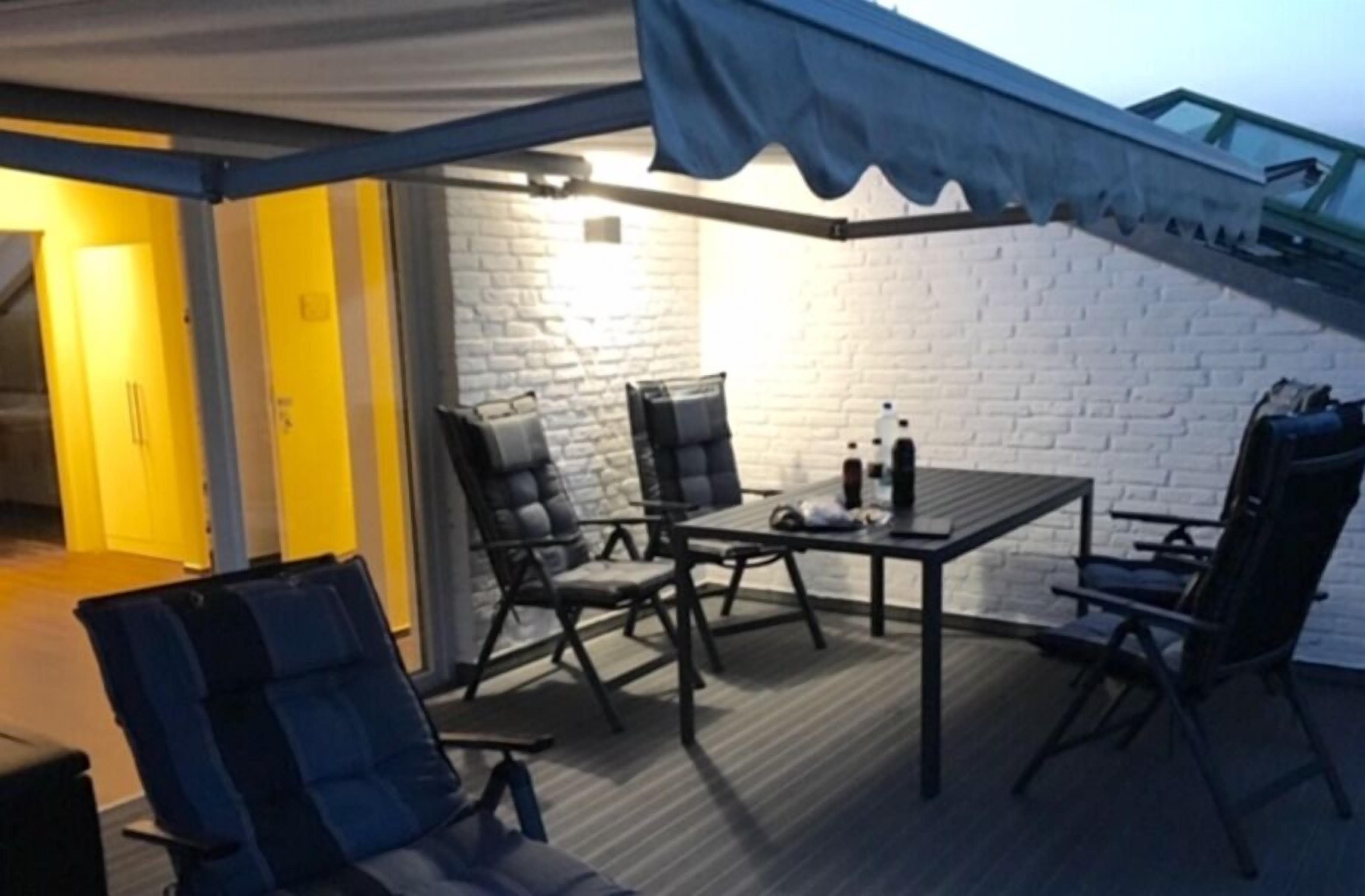 Una terraza en la azotea con una mesa y sillas, un toldo azul y un muro de ladrillo blanco.