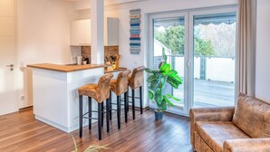 Een moderne keuken met een houten aanrecht en barstoelen, een zithoek met een bruine leren bank, en een grote schuifpui die uitkomt op een balkon met uitzicht op bomen.
