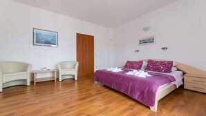 Ein Schlafzimmer mit Holzboden, einem Bett mit violetter Bettdecke, zwei Stühlen, einem kleinen Tisch und einer Tür.