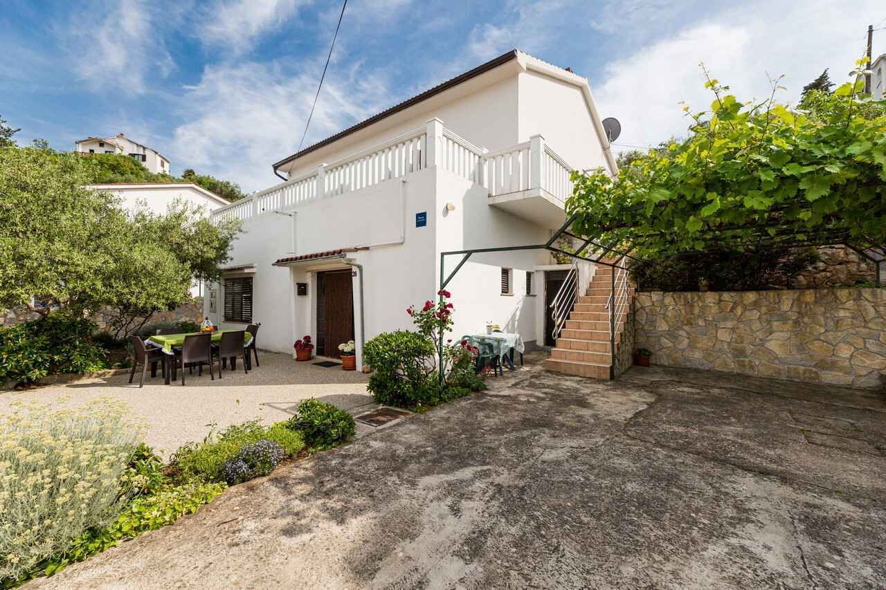 Una casa blanca de dos pisos con un balcón, un muro de piedra y un jardín con macetas y una mesa.