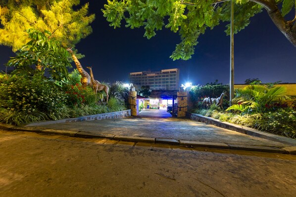 Vista nocturna de la entrada de un hotel, con un camino pavimentado que conduce a ella, rodeada de vegetación exuberante e iluminada por farolas.