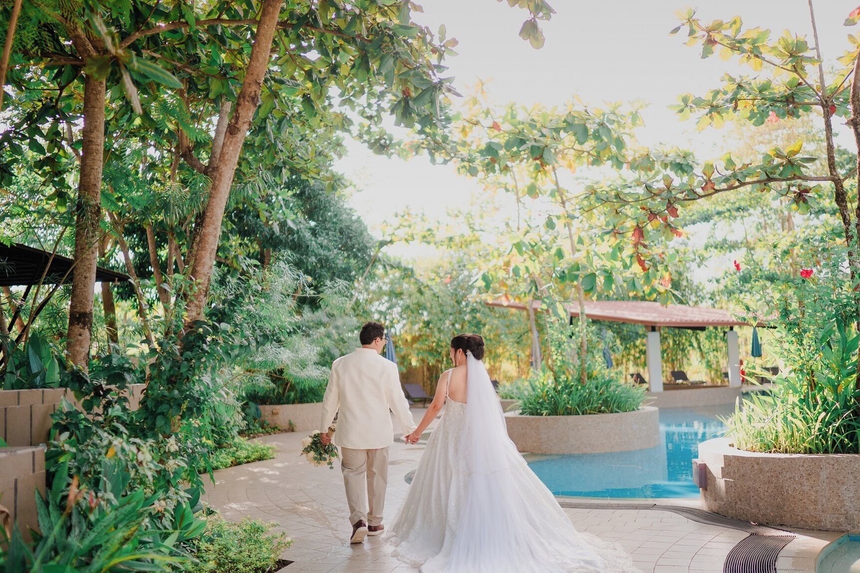A couple walking along a pathway with a pool and lush greenery in the background.