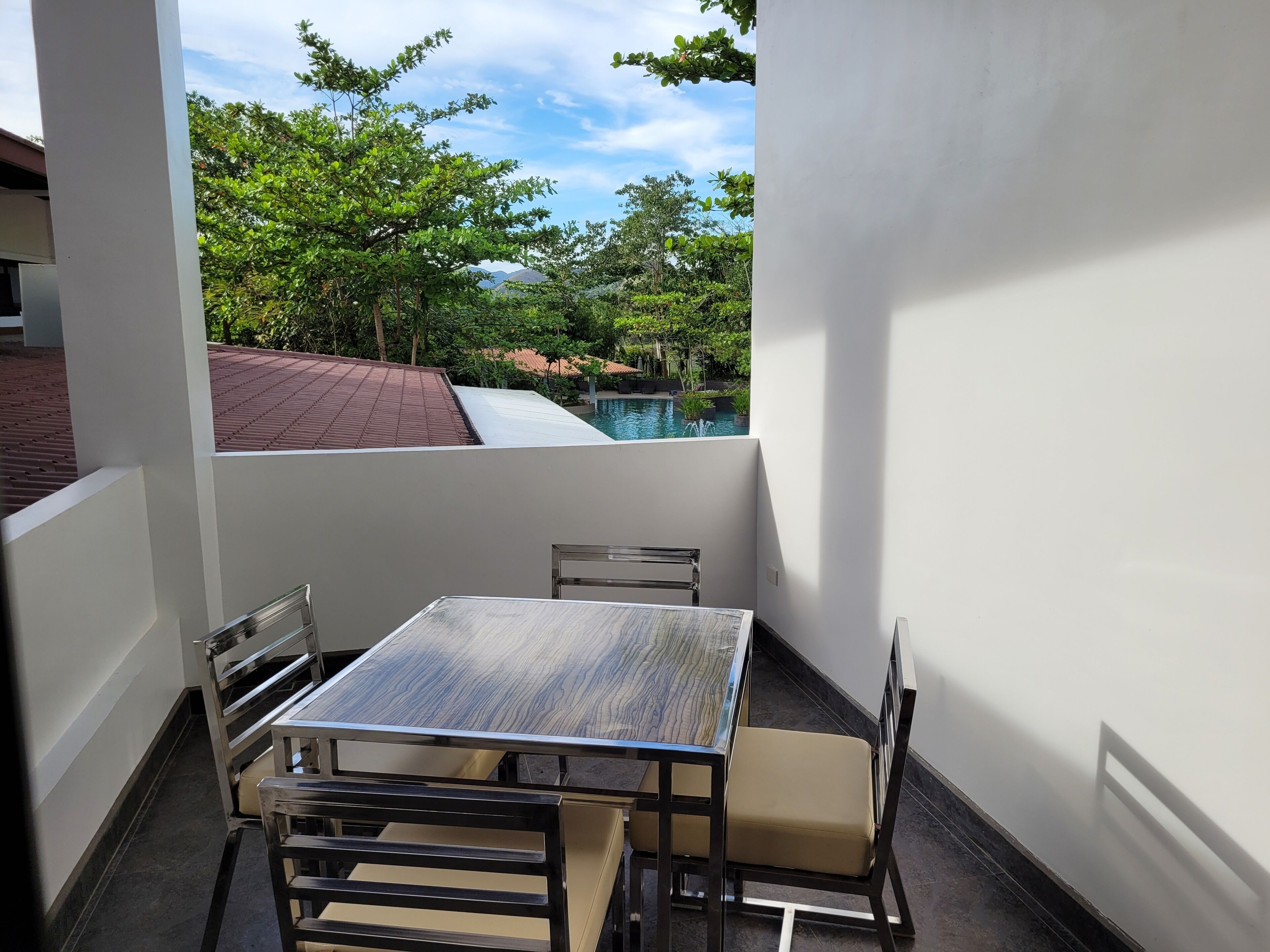 A small balcony with a table and chairs, overlooking a swimming pool and lush greenery.