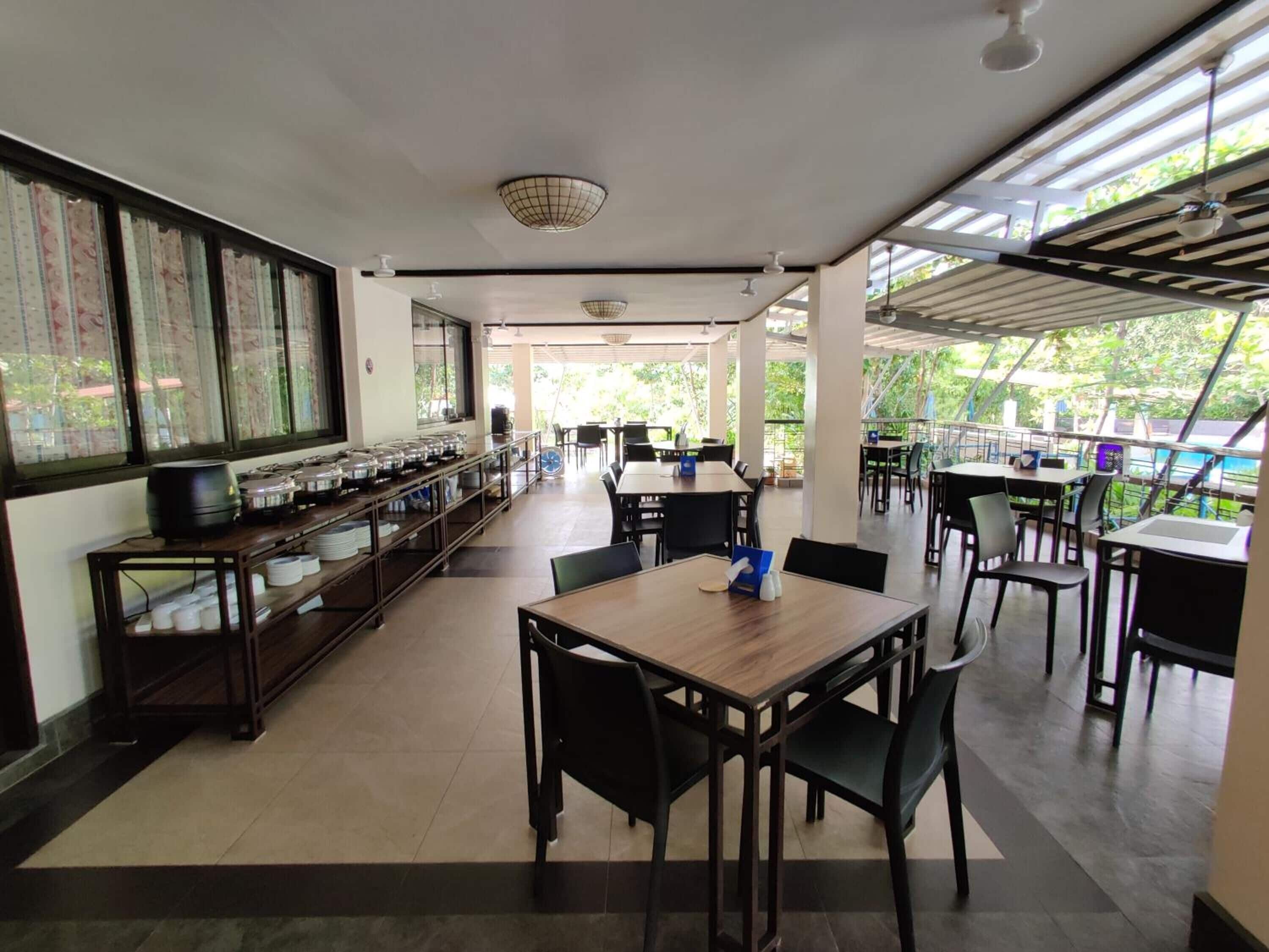 A spacious dining area with a long counter, multiple tables, and chairs. There are large windows providing natural light and a view of greenery outside.