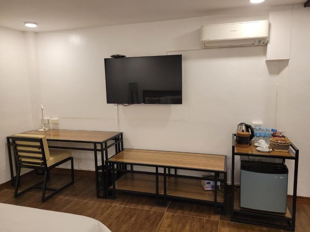 A compact hotel room with a bed, a desk with a chair, a television mounted on the wall, a small kitchenette, and an air conditioning unit.