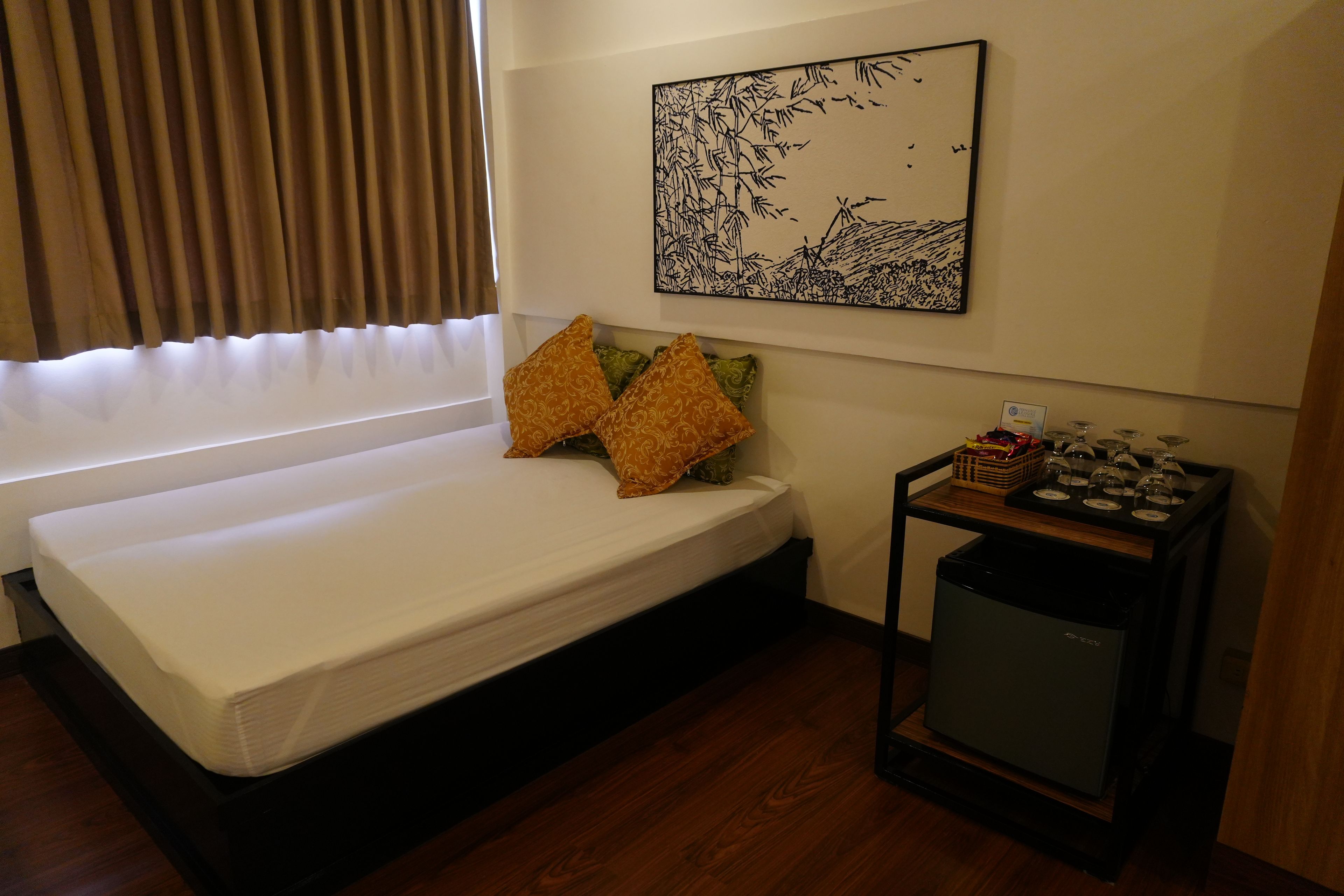 A small, neatly arranged room with a bed, a wall-mounted artwork, a small table with a basket, and a compact trash bin.