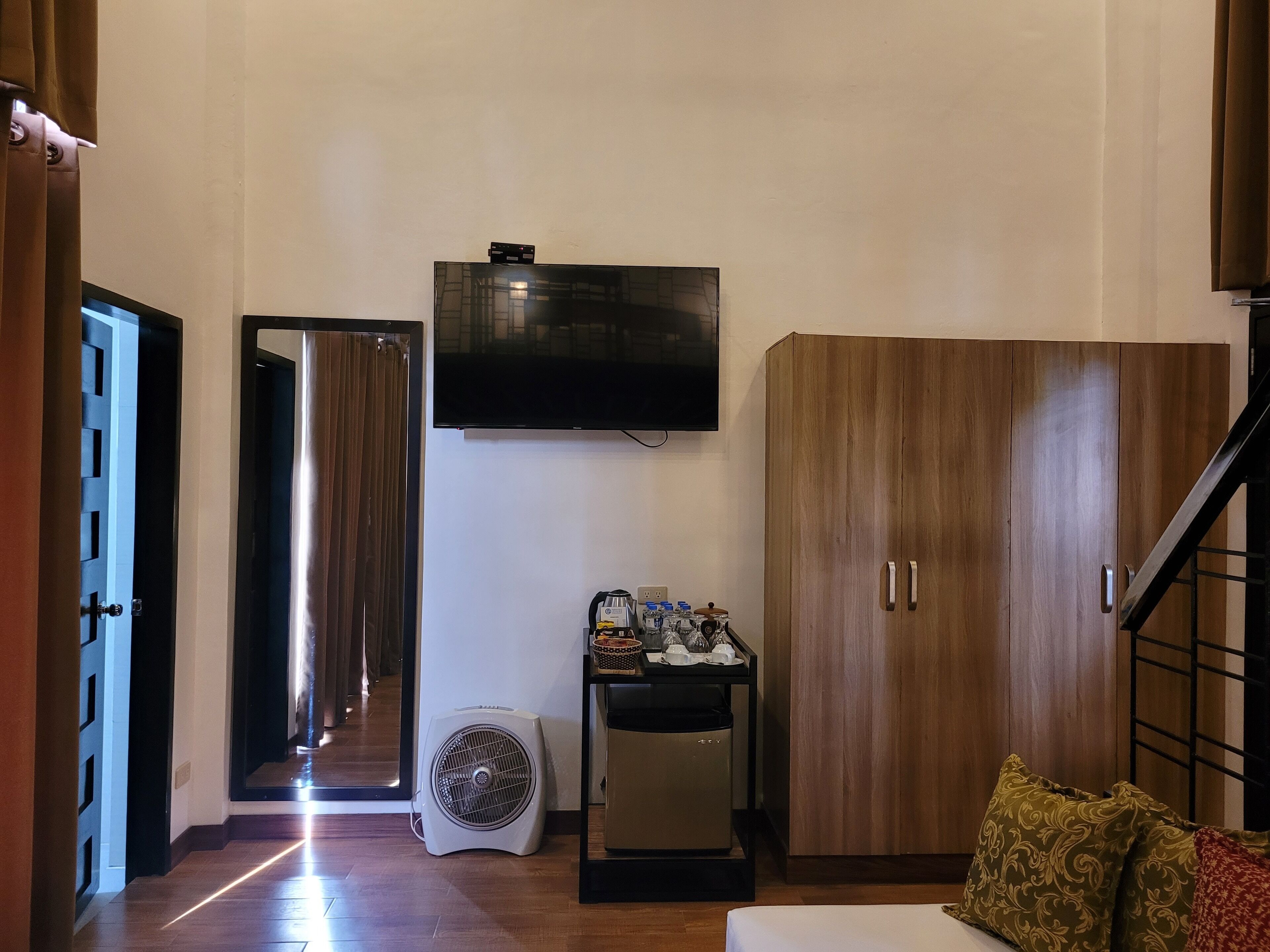 A hotel room with a TV mounted on the wall, a wardrobe, a small table with items, and a fan.