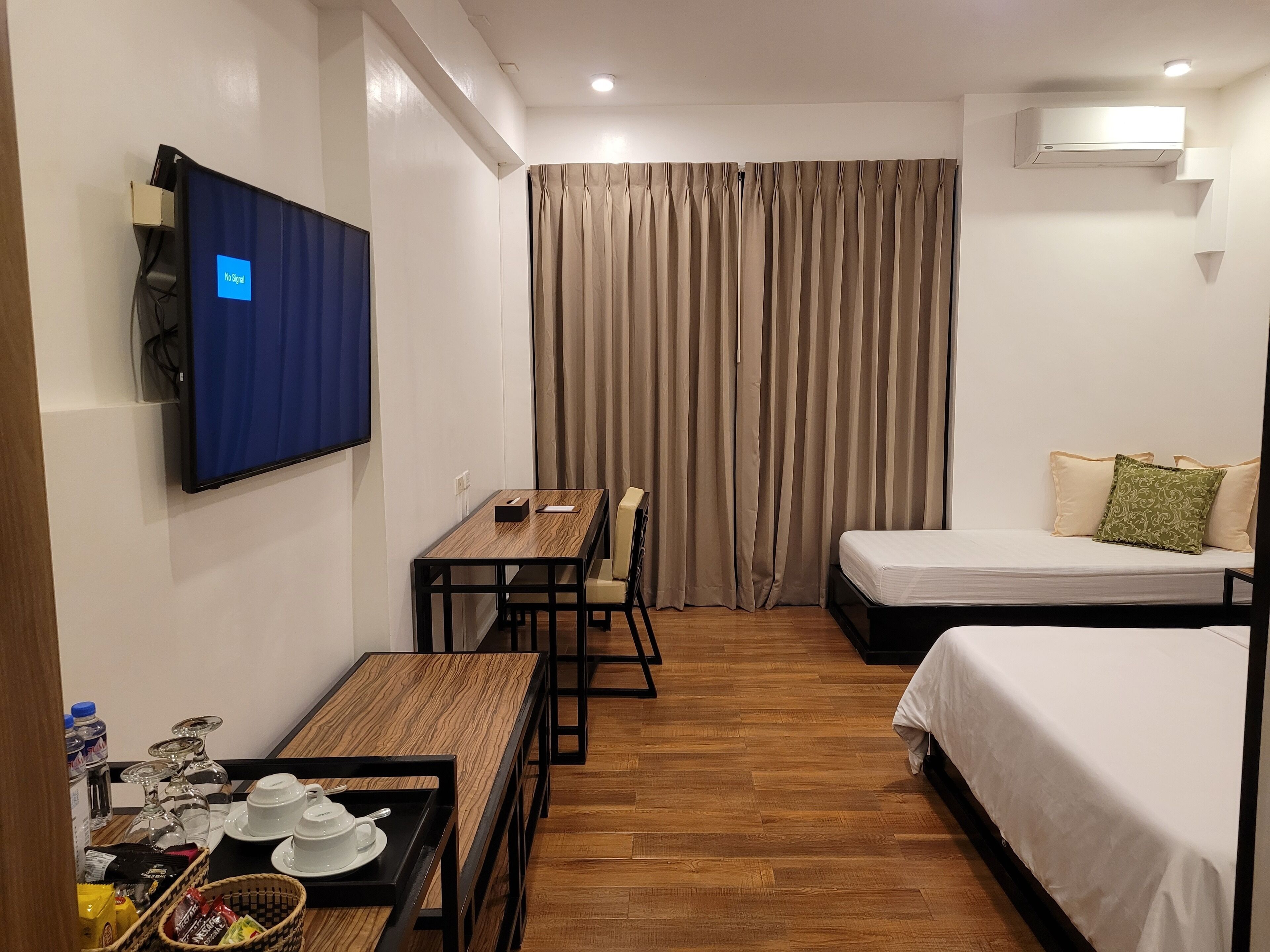 A hotel room with a wooden floor, a desk with a chair, a bed, a TV mounted on the wall, and a window with curtains.