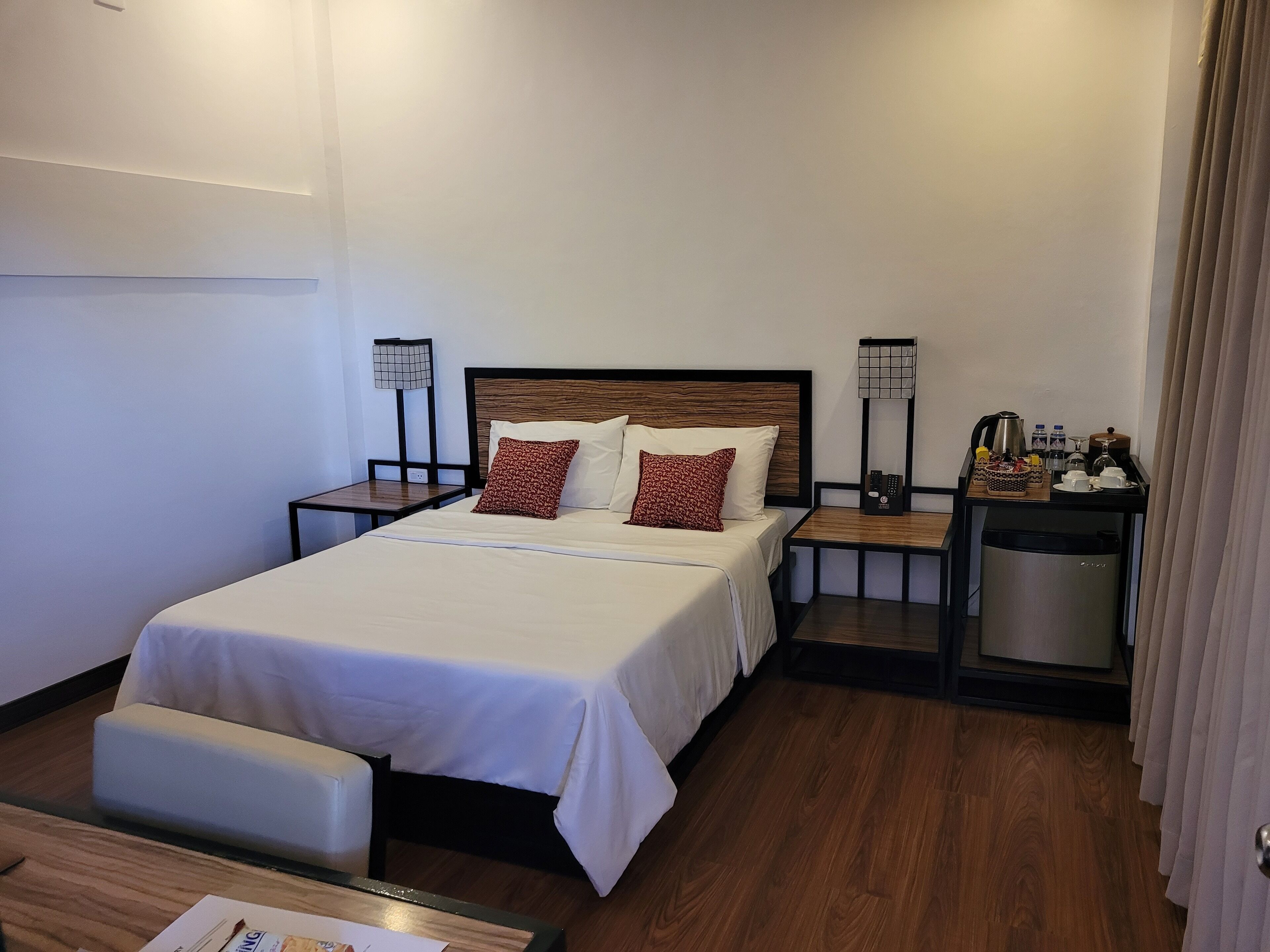 Una habitación de hotel con cama, mesitas de noche, lámpara, minibar y bandeja de desayuno.